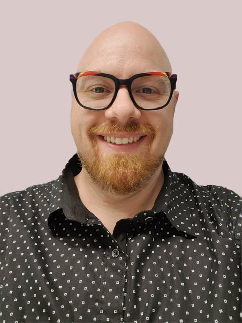Brian Young smiling, in rainbow-trimmed glasses and a black polka-dot shirt, on a soft pink-gray background